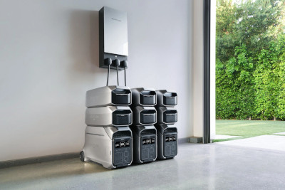 Three power stations, each with 2 extra batteries stacked on top, connect to a home power panel in a garage. The door is open with trees and grass in the background.