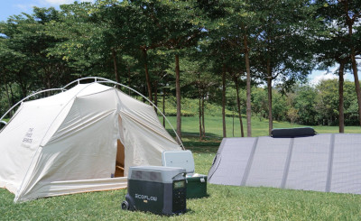 A solar panel charges a power station in front of a tent with a small portable refrigerator amid trees and grass.