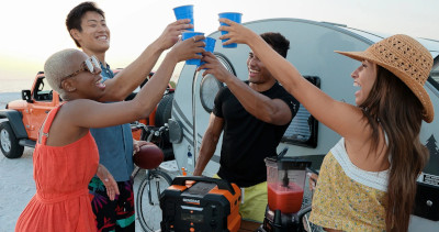 A group at a party on the beach toast their fun. A Generac Power Station power station provides power for a blender and a small camper towed by a jeep. You can see the beach sand and ocean in the background.
